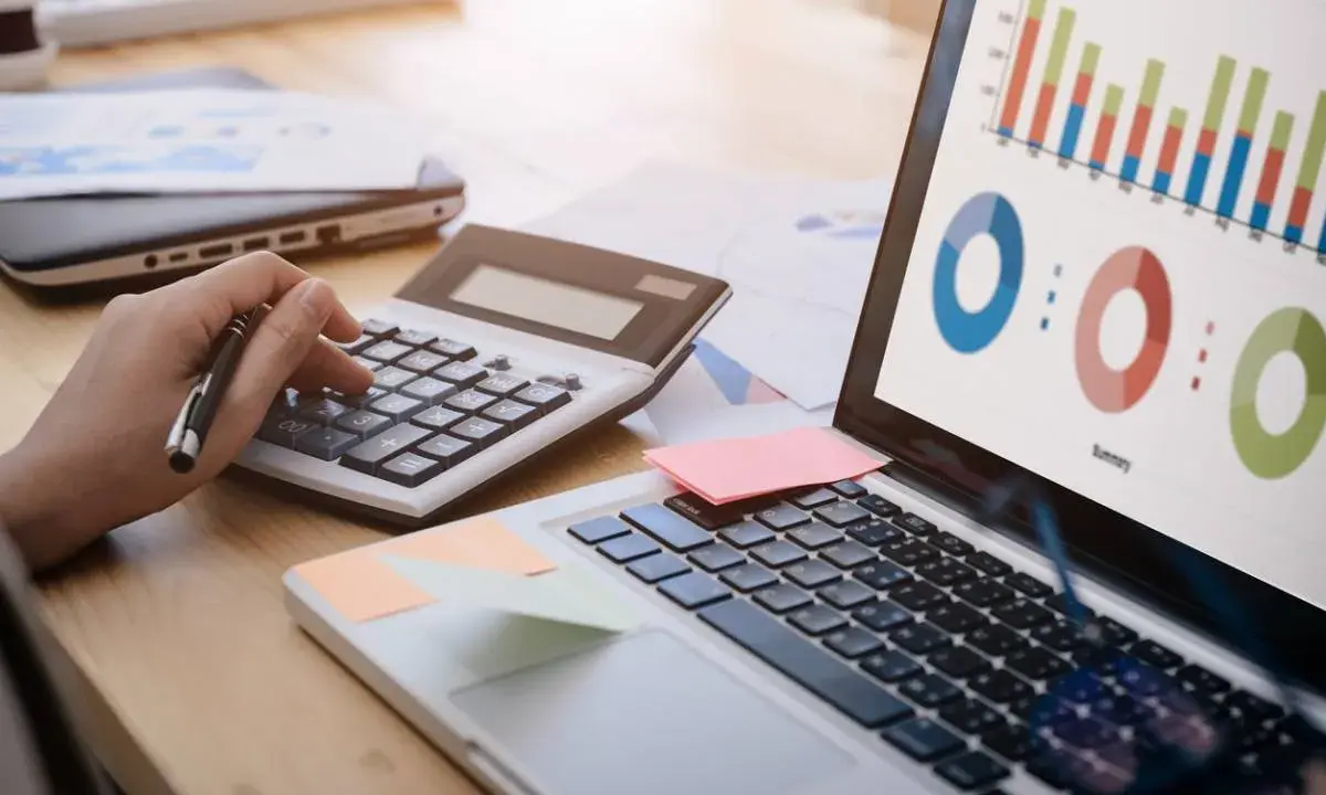 Main d'une personne utilisant une calculatrice pour le budget projet informatique, avec un ordinateur affichant des graphiques.