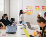 Une femme explique des techniques d'animation de réunion participative devant un mur couvert de post-its.