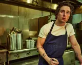 Un chef aux tatouages rêve d'ouvrir une pizzeria. Il est dans une cuisine professionnelle, entouré de casseroles et d'équipements.