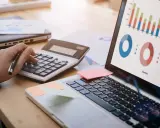 Main d'une personne utilisant une calculatrice pour le budget projet informatique, avec un ordinateur affichant des graphiques.
