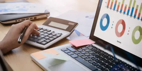 Main d'une personne utilisant une calculatrice pour le budget projet informatique, avec un ordinateur affichant des graphiques.
