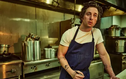 Un chef aux tatouages rêve d'ouvrir une pizzeria. Il est dans une cuisine professionnelle, entouré de casseroles et d'équipements.
