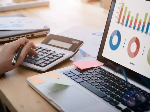 Main d'une personne utilisant une calculatrice pour le budget projet informatique, avec un ordinateur affichant des graphiques.