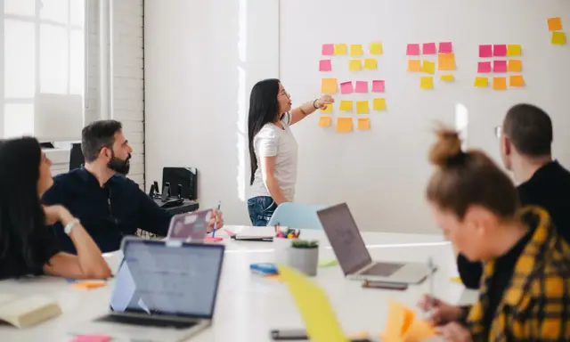Une femme explique des techniques d'animation de réunion participative devant un mur couvert de post-its.