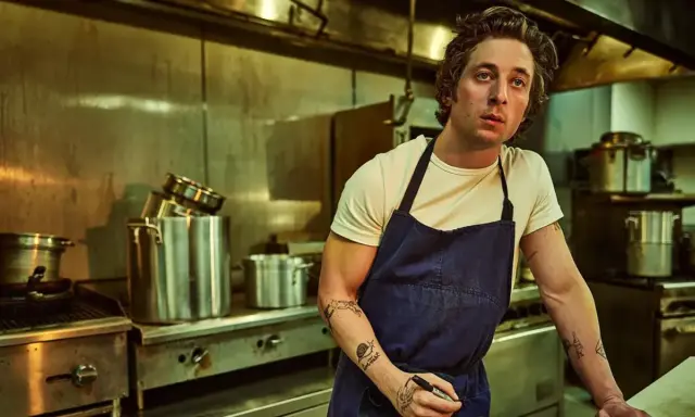 Un chef aux tatouages rêve d'ouvrir une pizzeria. Il est dans une cuisine professionnelle, entouré de casseroles et d'équipements.