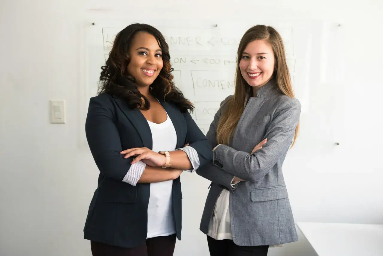 Deux femmes d'affaires souriantes, l'une noire et l'autre blanche, se tiennent devant un tableau blanc. La femme noire, qui pourrait être le chief of staff to the CEO, porte un blazer bleu marine.