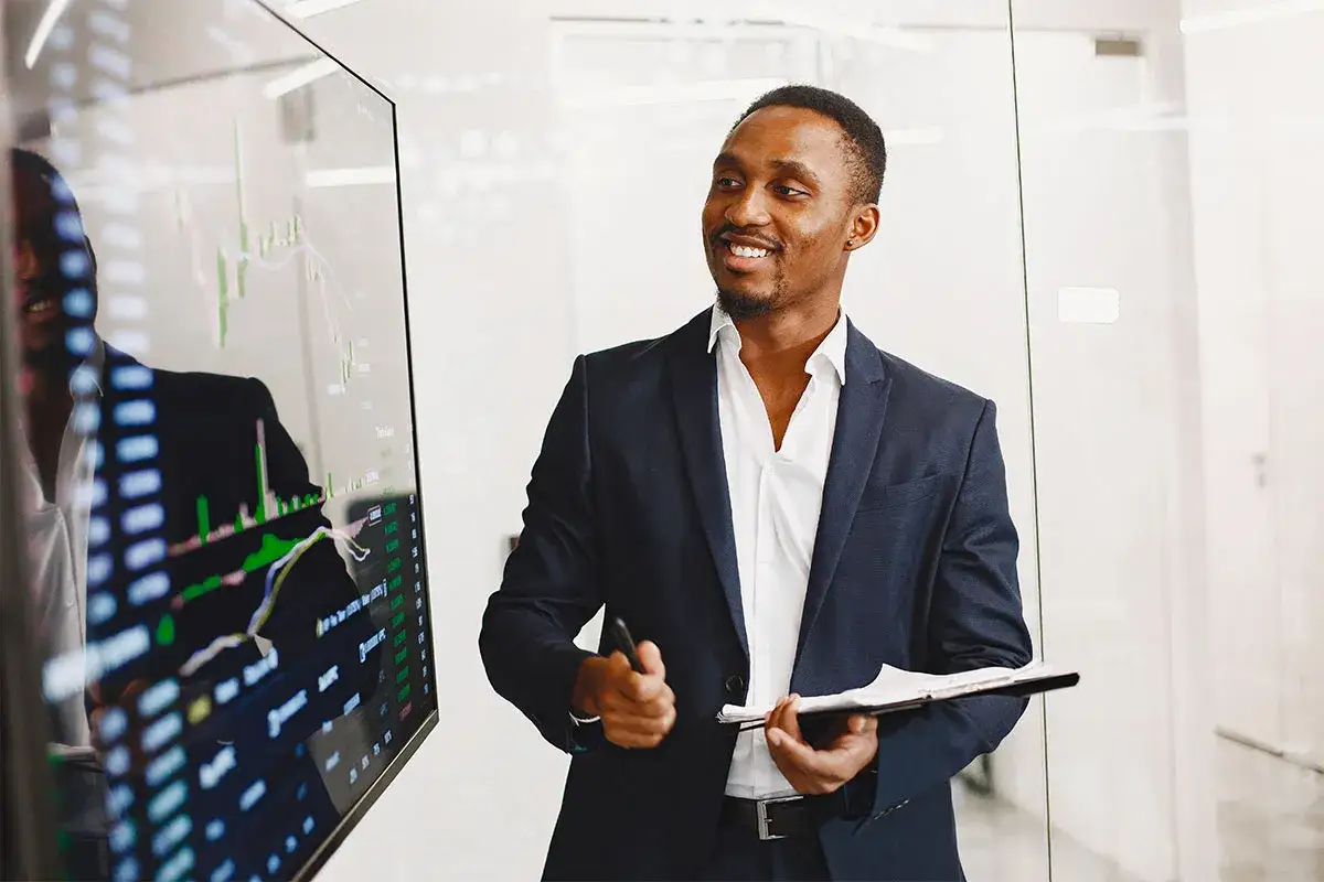 Un key account executive souriant pr&eacute;sente des graphiques financiers sur un &eacute;cran. Il tient un stylo et un bloc-notes.