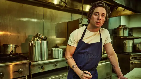 Un chef aux tatouages rêve d'ouvrir une pizzeria. Il est dans une cuisine professionnelle, entouré de casseroles et d'équipements.